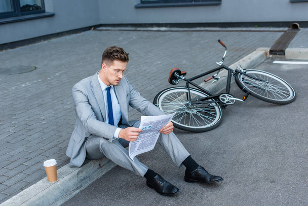 Businessman reading newspaper near coffee to go and bike on walkway  - Foto, Bild