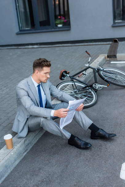 Businessman reading newspaper while sitting near takeaway coffee and bicycle outdoors  - Zdjęcie, obraz