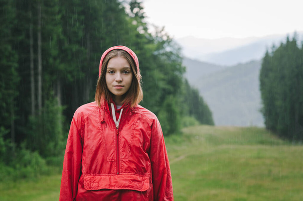 Portrait of a young beautiful lady standing on the mountain hill in the pouring rain wearing a red waterproof jacket, feeling peaceful and calm. - Foto, Imagem