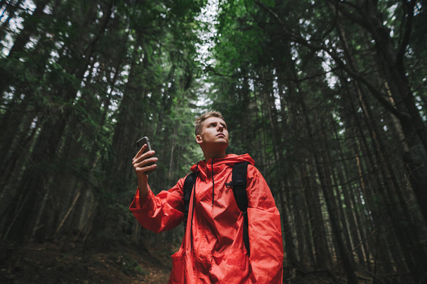 Bonito jovem usando seu telefone celular para encontrar sua localização na floresta de montanha, ele está procurando conexão com a internet. Cara vestindo um casaco de chuva vermelho e mochila. - Foto, Imagem