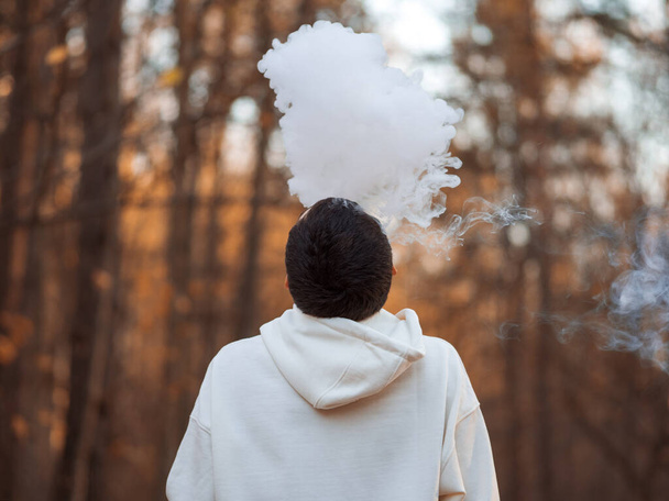 Een jongeman met een witte capuchon ademt een wolk stoomrook uit tegen de achtergrond van een herfstpark. - Foto, afbeelding