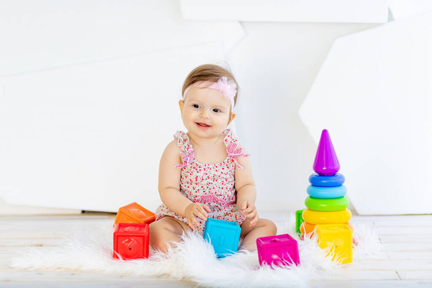 щаслива маленька дівчинка сидить у білій кімнаті в червоній сукні з яскравими іграшками
 - Фото, зображення