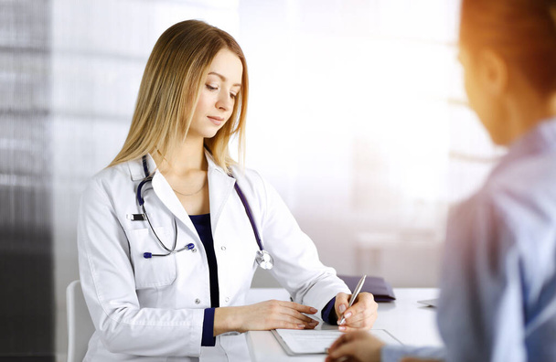 Young beautiful woman-doctor and her patient are discussing patients current health examination, while sitting together at the desk in the sunny cabinet in a clinic. Female physician is writing some - Фото, изображение