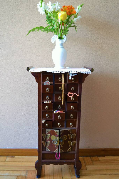 Комод китайського стилю з ювелірними виробами, що виступають зі своїх ящиків і вазою з квітами зверху
 - Фото, зображення