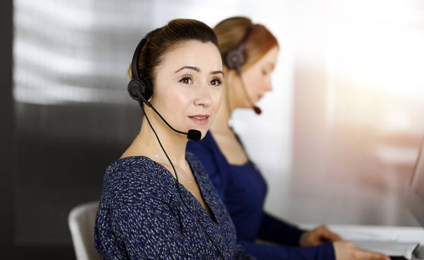 Two busineswomen have conversations with the clients by headsets, while sitting at the desk in a sunny modern office. Diverse people group in a call center. Telemarketing and customer service - Fotó, kép