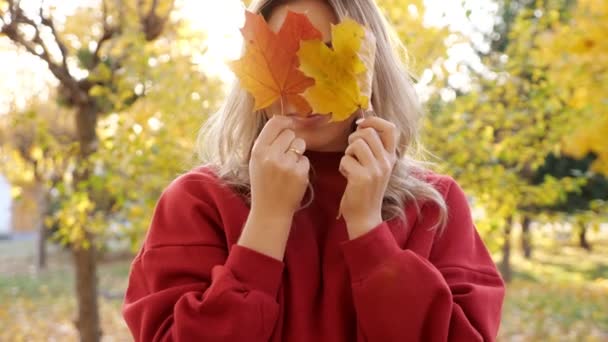 Vrouw in rode jurk verbergt gezicht met gele bladeren in park - Video