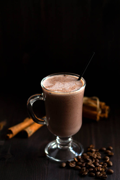 Ένα φλιτζάνι κακάο. Ένα ποτήρι ζεστή σοκολάτα με σαντιγί, κόκκους καφέ και ξυλάκια κανέλας σε σκούρο φόντο. ζεστά ροφήματα σε ξύλινο φόντο. Χειμώνας και φθινόπωρο. κατακόρυφα. αντίγραφο χώρου - Φωτογραφία, εικόνα