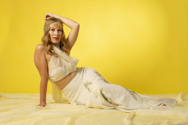 Mujer joven y bella vestida con falda, diadema dorada, y atillo de gasa sobre fondo amarillo, en sesion fotografica en estudio, diosa griega - Foto, Imagen