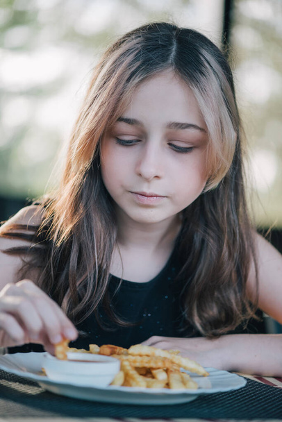 Belle petite fille riante assise à table et mangeant des frites de votre assiette. Adolescente mangeant des frites. heureuse petite fille manger un frites. - Photo, image