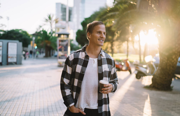 Iloinen valkoihoinen 20s mies matkustava nauttia vapaa-aikaa tutkia kaupungin sijainti ja määränpää lomilla, hymyilevä hipster kaveri trendikäs kulumista lepää kadulla aurinkoisena iltana cup - Valokuva, kuva