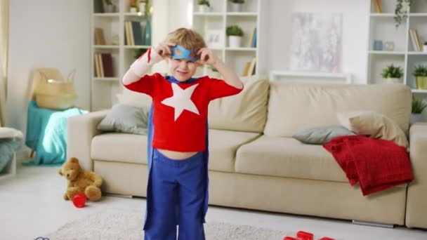 Střední záběr malého kudrnatého chlapce na rozmazaném pozadí v kostýmu superhrdiny s modrým pláštěm při pohledu na kameru a na sobě modrou oční masku napodobující létání s pravou paží a šklebení - Záběry, video