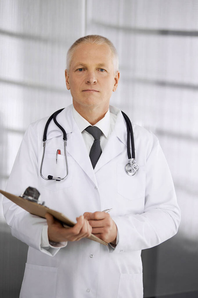 Elderly confident doctor standing straight and using clipboard in clinic. Perfect medical service. Medicine and healthcare concept - Photo, Image