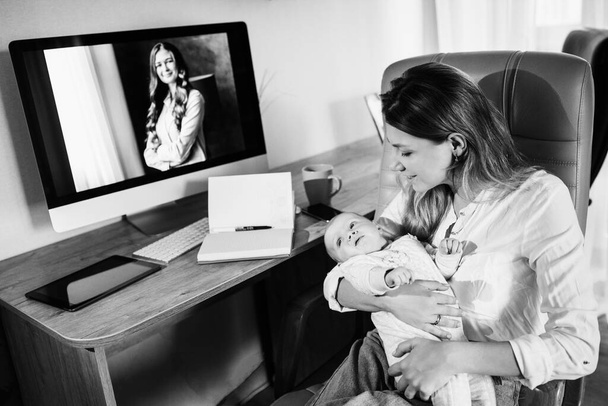 Happy young mother sit at the table, hold in arms little daughter, beautiful lady take a break with baby girl, smiling, business woman working at the home online - 写真・画像