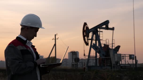 man in a white helmet with a laptop on the background of an oil rocking at sunset - Footage, Video