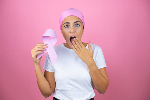 Young beautiful woman wearing pink headscarf holding brest cancer ribbon over isolated pink background cover mouth with hand shocked with shame for mistake, expression of fear, scared in silence, - Photo, Image