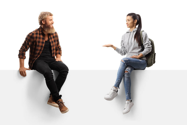 Bearded guy laughing and talking to a female student seated on a blank panel isolated on white background - Foto, immagini