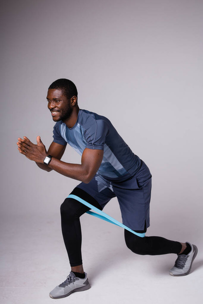 African american fitness trainer lunges with blue workout elastic. Shows exercise on a gray background - Foto, immagini