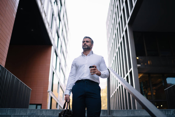 Pensive male entrepreneur with disposable takeaway cup and briefcase in hands feeling pondering at urban setting, middle aged businessman with coffee to go thinking about CEO during morning - Fotoğraf, Görsel