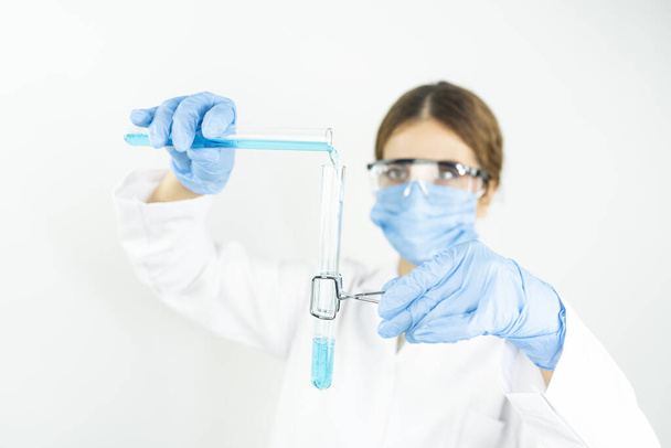 Joven científica femenina concentrada atractiva en anteojos protectores, máscara y guantes que dejan caer una sustancia líquida azul en el tubo de ensayo con una pipeta en el laboratorio químico científico    - Foto, Imagen