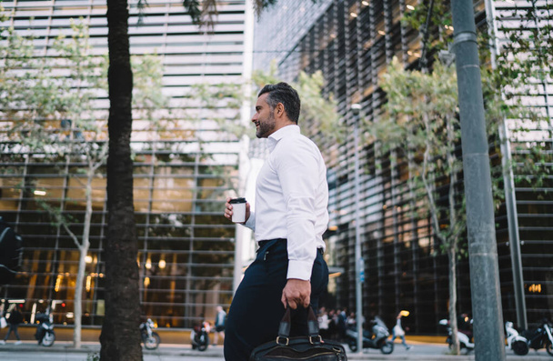 Side view of middle aged businessman with takeaway coffee to go and briefcase walking at urban street in financial district, Caucasian male entrepreneur dressed in white shirt holding disposable cup - Photo, Image
