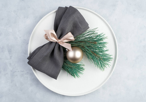 The white plate is decorated with a gray napkin with a pink bow, fir branches and a Christmas ball on a light concrete background. - Fotó, kép