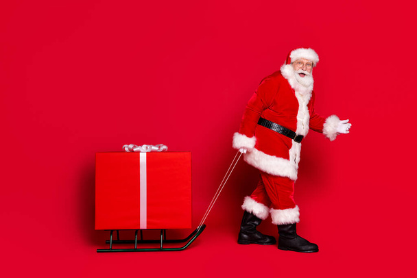 Oh no you found me. Photo portrait of astonished impressed stylish santa claus with white beard pulling big wrapped gift box on sleigh isolated on vivid red colored background - Фото, изображение
