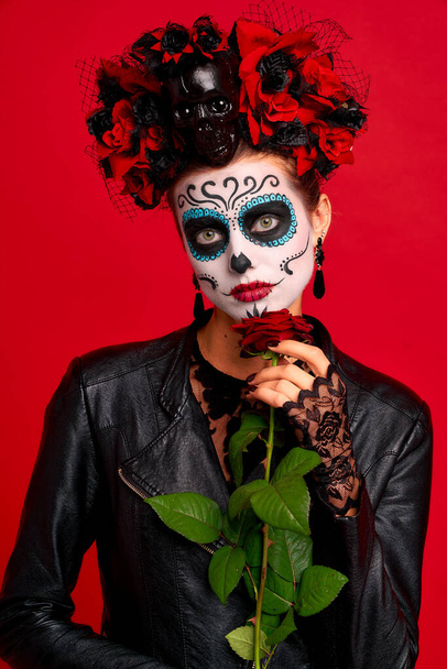 Romantic dead gir with sugar skull makeup with a wreath of flowers on her head and skull, wearth black gloves holding red rose isolated on red background. concept of Halloween or Calavera Catrina. - Photo, Image