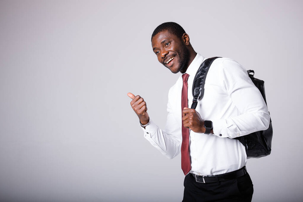 A male African American student in a white shirt, with a black backpack on his shoulder, stands on a gray background. Smiling, pointing left thumb - Zdjęcie, obraz