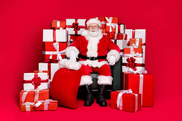Full length body size view of his he handsome fat overweight cheerful Santa Saint Nicholas sitting in chair pile stack giftboxes isolated bright vivid shine vibrant red color background - Zdjęcie, obraz