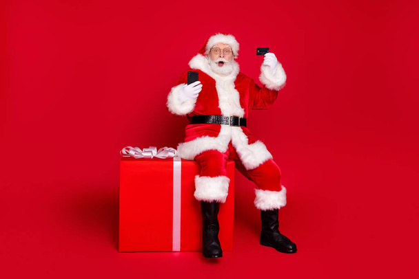 Full length body size view of his he handsome fat bearded amazed Santa sit on large big giftbox using device plastic card celebrate order shop isolated bright vivid shine vibrant red color background - Foto, immagini