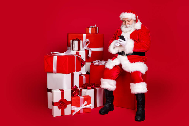 Full length body size view of his he nice handsome bearded focused Santa father sitting on big large giftbox using device shop order delivery isolated bright vivid shine vibrant red color background - Photo, image