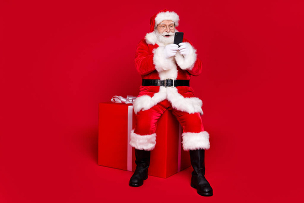 Повна довжина тіла погляд на його гарний красивий бородатий веселий батько Санта сидить на великій подарунковій коробці балачки веб-магазин доставки замовлення ізольовані яскравий яскравий блиск яскравий червоний колір фону
 - Фото, зображення
