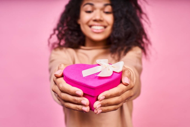 Young charming smiling girl with dark skin and curly curvy hair, wears a beige jacket, holds a bright pink box gift in the form of a heart with a white bow. - 写真・画像