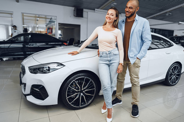 Happy couple of caucasian woman and african american man standing near their new car inside car salon - 写真・画像