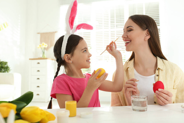 Happy daughter with bunny ears headband and her mother having fun while painting Easter eggs at home - Foto, Bild