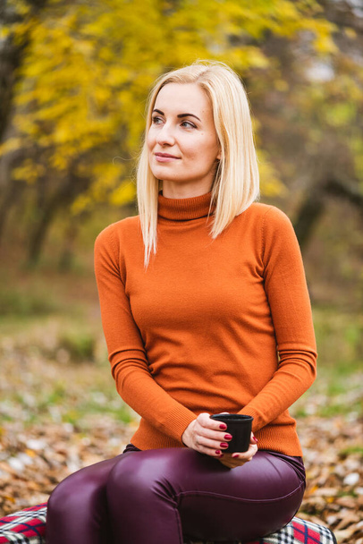 Pretty blonde young female with blue eyes in orange sweater hold black thermos cup with warming drink at yellow leaves background, bright fall season - Foto, afbeelding