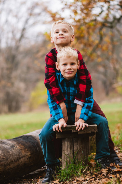 Zwei charmante Jungs im Grundschulalter von 6-7 Jahren sitzen an einer Holzbank, umarmen sich in ähnlich karierten Hemden vor herbstlichem Parkhintergrund. - Foto, Bild