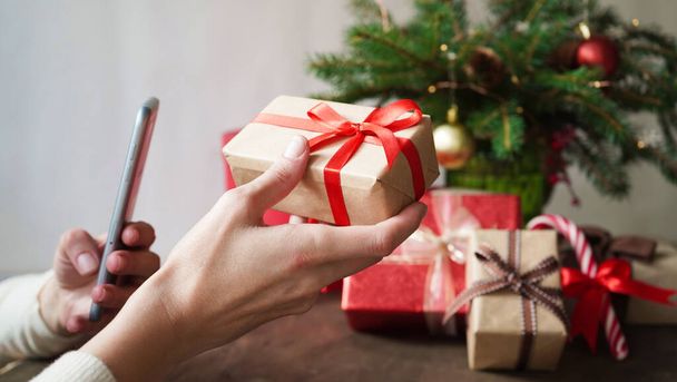 Elinde hediye kutusu ve akıllı telefon olan genç bir kadın Noel dekoru ve hediyelerin arka planında, yakın plan. İnternetten Noel ve Yeni Yıl alışverişi, kredi kartıyla ödeme. - Fotoğraf, Görsel