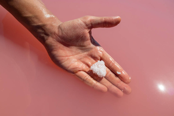 Χέρι κρατώντας λευκό κρύσταλλο αλάτι σε ροζ ζωντανό αλμυρό νερό ηλιόλουστο γκρο πλαν. Spa διαδικασίες στο φυσικό θέρετρο - Φωτογραφία, εικόνα
