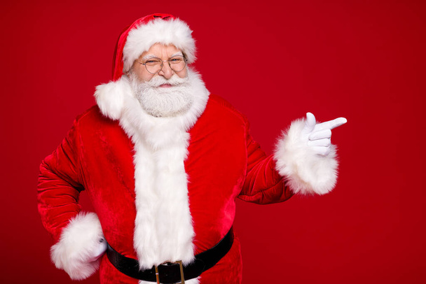 Photo of retired old man grey beard hand hip direct finger empty space propose newyear decor wear santa costume leather belt gloves coat spectacles headwear isolated red color background - 写真・画像