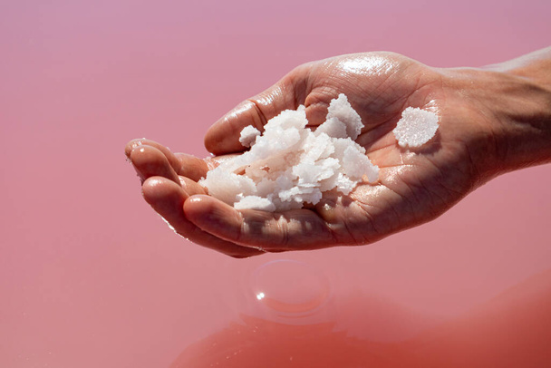 Χέρι κρατώντας δέσμη ροζ λευκό αλάτι νιφάδες κρυστάλλους πάνω από ροζ ζωντανή επιφάνεια του νερού της λίμνης. Spa θέρετρο ηλιόλουστη close-up - Φωτογραφία, εικόνα