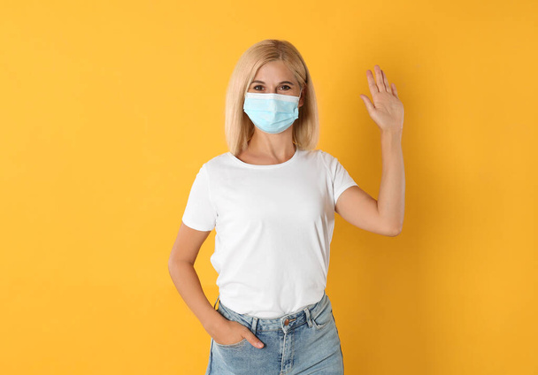 Woman in protective mask showing hello gesture on yellow background. Keeping social distance during coronavirus pandemic - Photo, Image