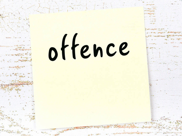 Yellow sticky note on wooden wall with handwritten word offence - Photo, Image