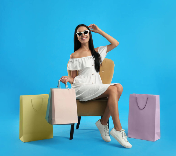 Beautiful young woman with paper shopping bags in armchair on light blue background - Fotó, kép