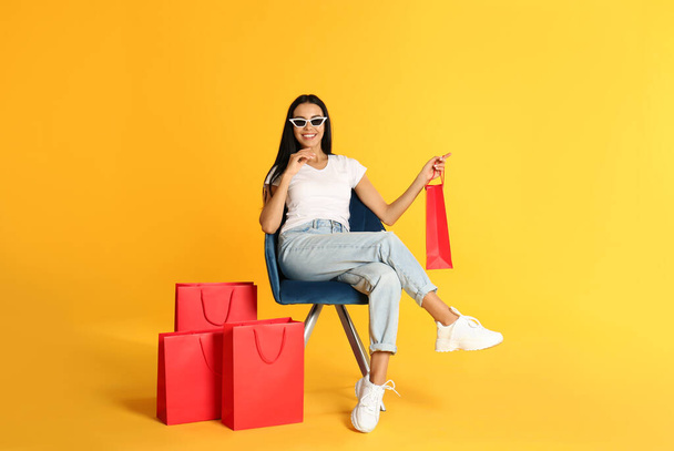 Beautiful young woman with paper shopping bags in armchair on yellow background - Foto, Imagem