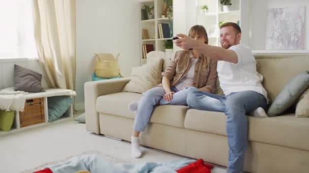 Handheld shot van ouders glimlachen en praten in de voorkant van TV, terwijl jonge zoon met teddybeer en dochter in superheld kostuums met oogmaskers slapen op de vloer bedekt met deken in de woonkamer - Video