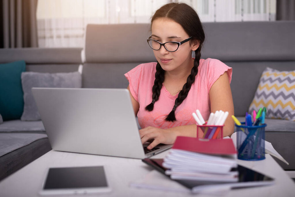 社会的距離のある学生の概念、家庭での教育。高校生の女の子はノートパソコンを持って家で宿題をしている。ティーンスタディホームザ学校が閉鎖中コロナウイルスCovid-19. - 写真・画像