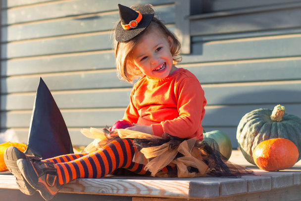 Słodka dziewczynka 2-3 w pomarańczowym i czarnym kostiumie czarownicy Halloween, siedzi obok dyń na tarasie drewnianej szarej kamienicy - Zdjęcie, obraz