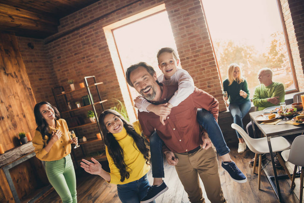 Photo of full family gathering six people two small kids play dad piggyback daughter wave hand mom laugh grandparents sit sofa dinner table generation in home evening living room indoors - Fotó, kép
