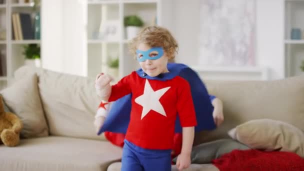 Střední záběr mladého chlapce v kostýmu superhrdiny s modrou oční maskou napodobující létání jako hrdina s pěstmi vpřed a starší sestra sedí na pohovce mává pláštěm v obývacím pokoji - Záběry, video